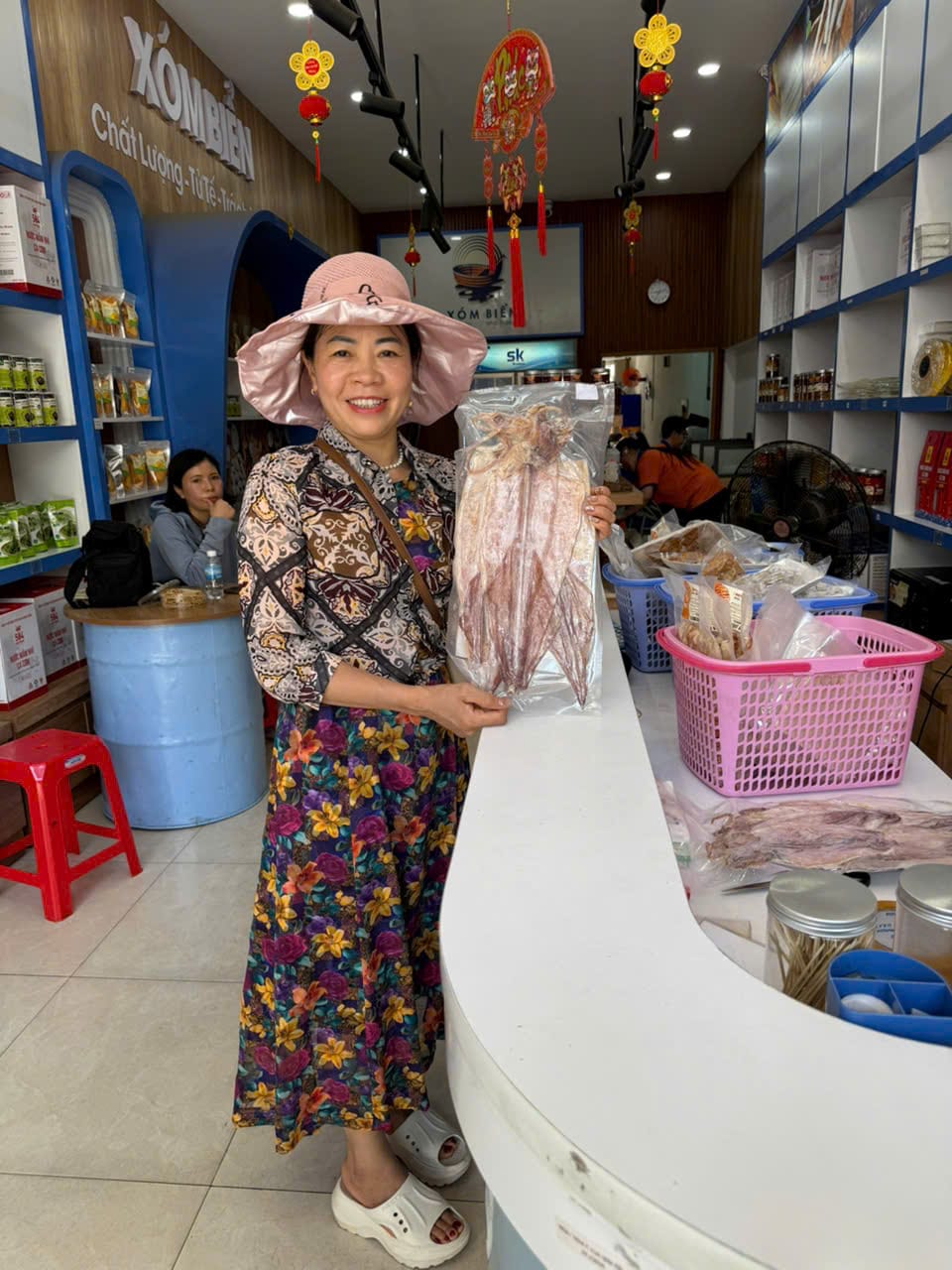Mực Khô Nha Trang Khác Mực Một Nắng Thế Nào? Loại Nào Nên Mua 4 Khách hàng cầm túi mực khô nguyên con, cười tươi trước bảng hiệu Xóm Biển